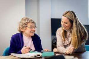 Two women, one older, one slightly younger, sitting side by side at a table, in discussion